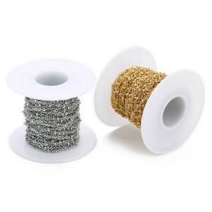 May include: Two spools of chain, one gold and one silver, are wound around white plastic spools. The spools are on a white background.