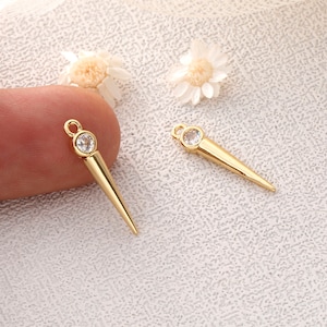 Pode incluir: Dois pingentes de joia dourados em forma de espinho, com uma gema redonda e transparente no topo. Cada pingente tem uma pequena argola para prender a uma corrente ou outro componente de joia. Os pingentes são exibidos em uma superfície texturizada.