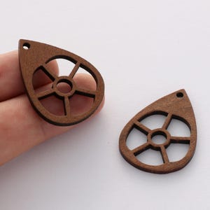 May include: Two brown wooden teardrop-shaped pendants with a circular cutout design. The pendants have a hole at the top for hanging.