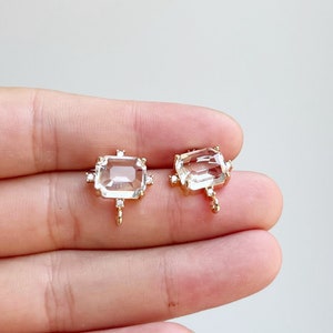 Pode incluir: Dois pingentes dourados com pedras preciosas facetadas transparentes rodeadas por pedras preciosas transparentes menores. Cada pingente tem um pequeno laço na parte inferior.