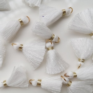 May include: A collection of white tassel trims with gold accents. The tassels are arranged on a white surface.