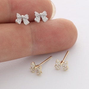 Peut inclure: Deux paires de boucles d'oreilles à tige en forme de nœud, une paire en métal argenté et une paire en métal doré. Chaque boucle est incrustée de petites pierres claires et scintillantes et possède une tige à l'arrière pour la fixation.