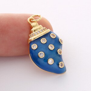 May include: A gold-colored pendant in the shape of a blue seashell. The pendant is adorned with clear, round gemstones and a ring for attachment. The seashell has a glossy finish and is held by a finger.
