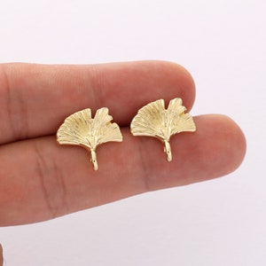 Può includere: Un paio di orecchini dorati con un design a foglia di ginkgo. Le foglie sono dettagliate con venature testurizzate e hanno un piccolo anello nella parte inferiore per fissarle a un perno.