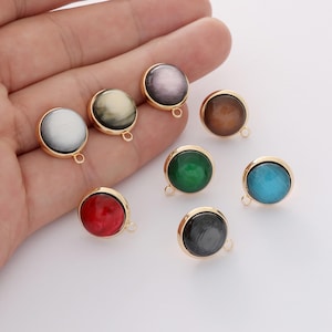 May include: A collection of eight circular cabochon charms, each featuring a smooth, chatoyant stone set in a gold-toned metal bezel. The charms display a variety of colors including white, cream, purple, brown, green, blue, red, and dark gray.