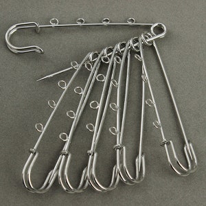 May include: A set of seven silver safety pins with multiple loops on each pin. The pins are arranged in a fan shape.