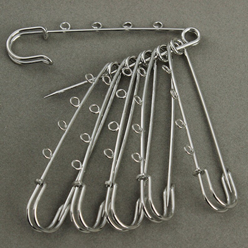 May include: A set of seven silver safety pins with multiple loops on each pin. The pins are arranged in a fan shape.