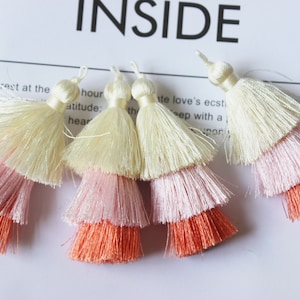 May include: Four multi-colored tassels with three layers of fringe. The top layer is white, the middle layer is pink, and the bottom layer is peach.