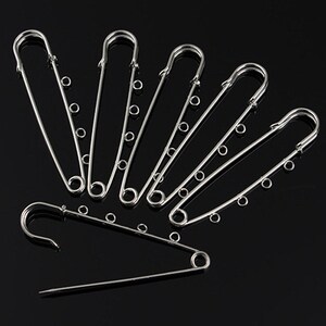 May include: A set of six silver safety pins with a chain detail. The pins are arranged in a fan shape on a black background.