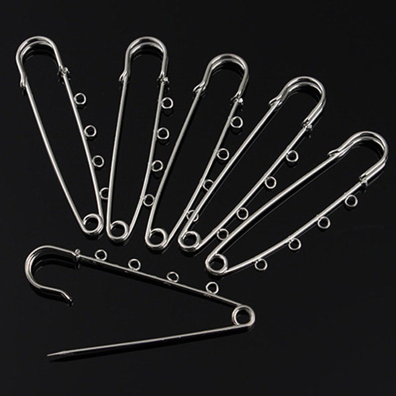 May include: A set of six silver safety pins with a chain detail. The pins are arranged in a fan shape on a black background.