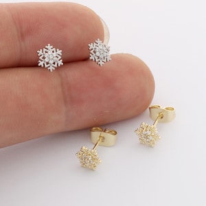 May include: Two pairs of snowflake stud earrings, one pair in silver-tone metal and one pair in gold-tone metal. Each earring features a central cluster of small clear stones arranged in a snowflake shape with six points and a post backing.
