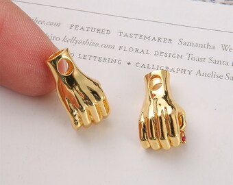 May include: Two gold-colored metal hand charms with a hole for stringing. One charm has a red stone on the finger.