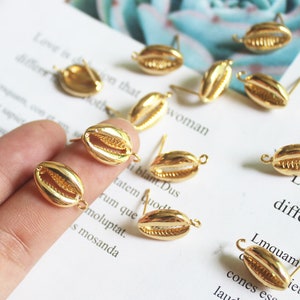 Könnte beinhalten: Eine Sammlung goldfarbener Muschel-Anhänger mit Sprungringen. Die Anhänger sind auf einer weißen Oberfläche mit Text im Hintergrund angeordnet.