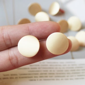May include: Two gold-colored round metal earrings, held in a hand, against a blurred background.