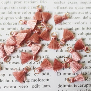 May include: A collection of small, dusty rose-colored tassels with gold-colored metal loops. These decorative tassels are made of soft, silky threads and are ideal for jewelry making or craft projects.