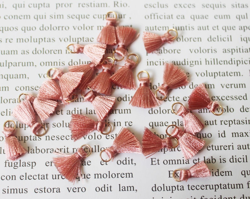 May include: A collection of small, dusty rose-colored tassels with gold-colored metal loops. These decorative tassels are made of soft, silky threads and are ideal for jewelry making or craft projects.