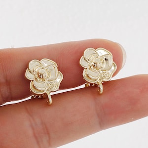 Puede incluir: Un par de pendientes de clip de tono dorado con un diseño floral. Los pendientes tienen forma de rosa con un centro elevado y una superficie texturizada.