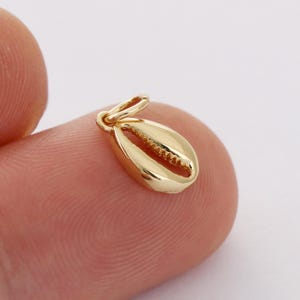 Könnte beinhalten: Goldfarbener Muschel-Anhänger. Der Anhänger ist in Form einer Kaurischnecke geformt und hat oben eine Öse zum Anbringen an einer Halskette oder einem Armband.