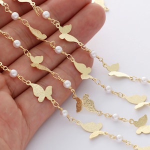 May include: Gold chain with white pearl beads and butterfly charms. The chain is on a hand, showing the detail of the charms.