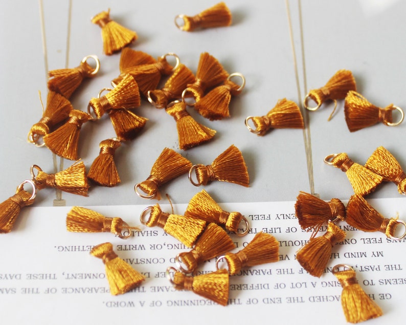 May include: A collection of small, gold-colored tassels with a loop at the top. The tassels are made of a soft, silky material and are perfect for adding a touch of elegance to any project.