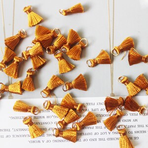 May include: A group of 20 mustard yellow tassel trim with gold metal rings. The tassels are made of soft, fine threads and are approximately 1 inch in length.