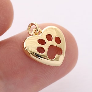 May include: A gold-colored heart-shaped pendant with a paw print cutout. The pendant has a loop at the top for a chain. The paw print design features three small circles and one larger oval shape.