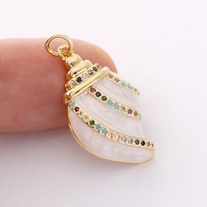 May include: A gold-colored seashell pendant with white enamel and colorful gemstones. The pendant has a loop at the top for attaching to a chain or bracelet. The gemstones are in various colors, including blue, red, and green.