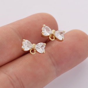 Puede incluir: Un par de pendientes dorados en forma de lazo con piedras de circonita cúbica claras y brillantes. Los pendientes tienen un diseño de poste y bucle para sujetarlos a la joyería. El diseño del lazo es un estilo clásico y elegante.