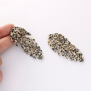May include: A pair of long, dangling earrings featuring a layered leaf design. The leaves are beige with black spots, giving them a leopard print look.