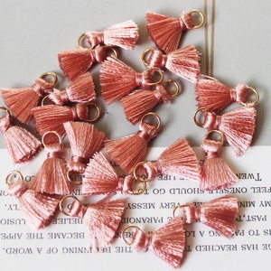 May include: A collection of small, peach-colored tassels with gold-colored metal loops. The tassels are arranged in a scattered pattern, with each tassel featuring a looped top and a fringed bottom. These are ideal for jewelry making or craft projects.