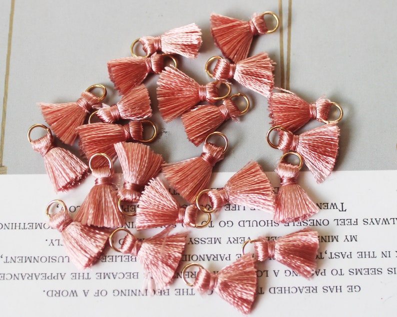 May include: A collection of small, peach-colored tassels with gold-colored metal loops. The tassels are arranged in a scattered pattern, with each tassel featuring a looped top and a fringed bottom. These are ideal for jewelry making or craft projects.