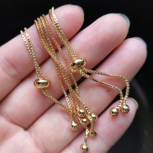 May include: A gold chain bracelet with multiple strands and adjustable sliding beads. The bracelet is made of a delicate chain and has a simple, elegant design.