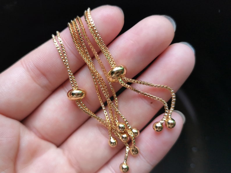 May include: A gold chain bracelet with multiple strands and adjustable sliding beads. The bracelet is made of a delicate chain and has a simple, elegant design.