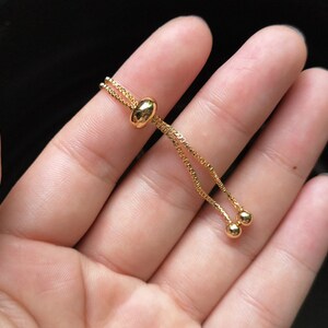 May include: A gold adjustable bracelet with a sliding bead closure. The bracelet is made of a delicate chain with two small round beads at the end.