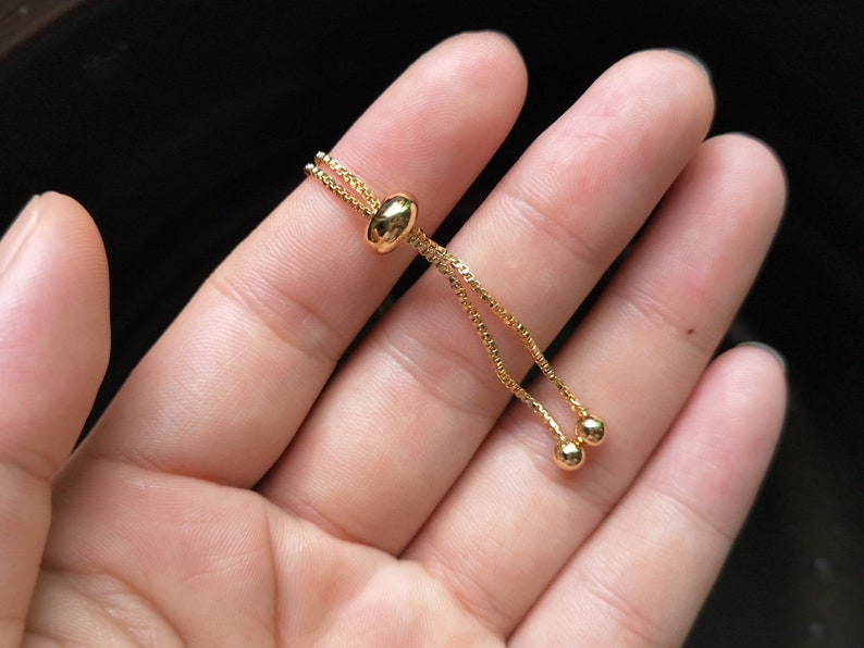May include: A gold adjustable bracelet with a sliding bead closure. The bracelet is made of a delicate chain with two small round beads at the end.