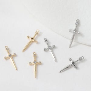 May include: An assortment of small sword-shaped charms. Some are gold-toned, others silver-toned, each featuring a decorative hilt with clear, round gemstones. The charms are designed for jewelry making or crafting.
