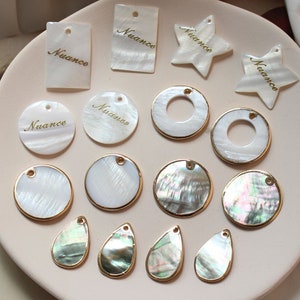 May include: Assortment of white and iridescent shell charms with gold accents. The charms are in various shapes, including circles, ovals, stars, and rectangles. Some charms have the word "Nuance" written on them.