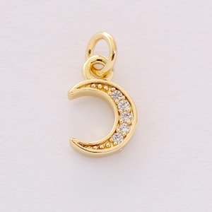 May include: A gold-colored crescent moon charm with a loop for hanging. The charm is embellished with small, clear gemstones along the inner curve and tiny gold beads along the outer edge. The charm is set against a white background.