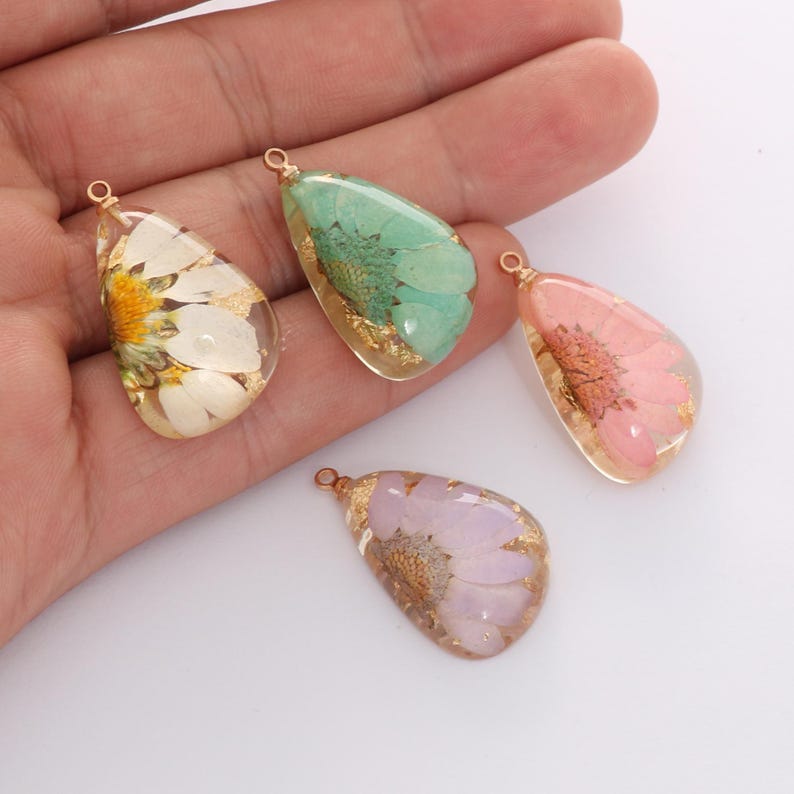 May include: Four teardrop-shaped resin pendants with dried flowers and gold flakes. The pendants feature a white and yellow daisy, a green flower, a pink flower, and a purple flower. Each pendant has a gold-colored loop at the top.