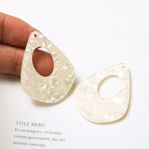 May include: Two pear-shaped, off-white acrylic earring pendants with a marbled pattern and a large oval cutout. Each pendant has a small hole at the top for attachment. The text "TITLE HERE" is visible in the background.