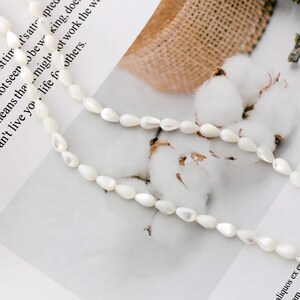 May include: A strand of white pearlescent teardrop-shaped beads, strung on a white cord. The beads are arranged in a loose, flowing pattern, and the cord is visible between the beads.