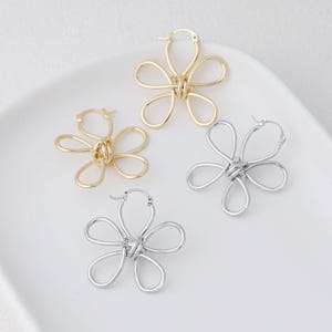 Puede incluir: Cuatro pendientes de aro con forma de flor en tonos dorados y plateados. Los pendientes están colocados sobre una superficie blanca. Cada pendiente presenta cinco pétalos en bucle conectados en el centro, creando un diseño floral.