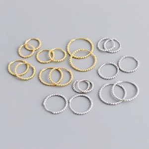 May include: A set of silver and gold hoop earrings with a beaded design. The earrings are of various sizes and are arranged in a symmetrical pattern on a grey background.