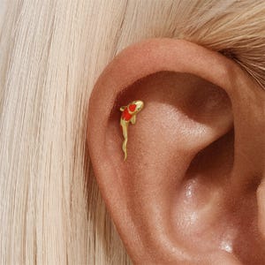 May include: A gold-coloured koi fish earring with red and white details is shown on an ear. The fish has a small, clear gem near its head. The earring is a unique piece of jewellery.