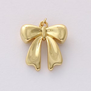 Op de afbeelding: Een gouden metalen strikbedel met een lus aan de bovenkant om aan sieraden of andere knutselwerkjes te bevestigen.