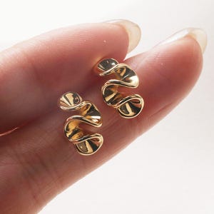 Puede incluir: Un par de pendientes de color dorado con un diseño ondulado y orgánico. Los pendientes son pequeños y delicados, y serían una gran adición a cualquier colección de joyas.