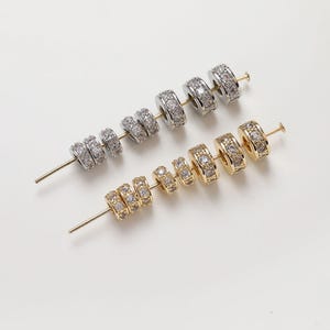 May include: Two strands of jewelry beads with clear rhinestones. One strand is silver and the other is gold. The beads are round and have a raised edge.