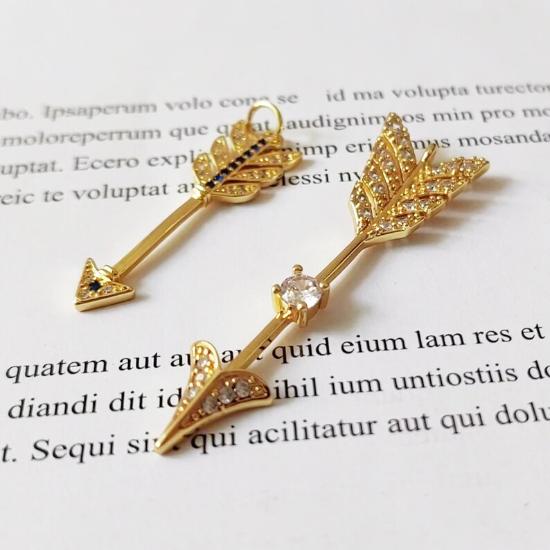 May include: Two gold arrow earrings with clear and black crystal accents. The arrowheads are adorned with small crystals, and the shafts are embellished with larger crystals.