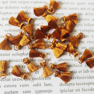 May include: A collection of 20 small, mustard yellow tassel charms with gold jump rings. The tassels are made of soft, fuzzy yarn and are approximately 1 inch long.