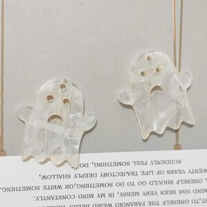 Puede incluir: Dos adornos blancos en forma de fantasma con detalles de purpurina. Los fantasmas tienen una cara triste con tres agujeros para los ojos y una boca.
